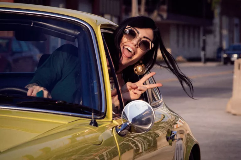 A woman wearing sunglasses flashes a peace sign out the window of a yellow sports car