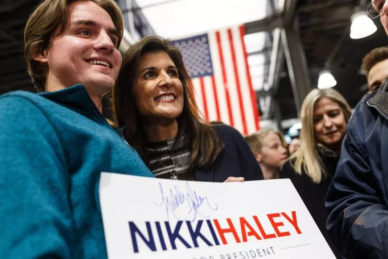 Former UN Ambassador Nikki Haley poses for a photo with a supporter holding a campaign placard. A large American flag hangs in the background.